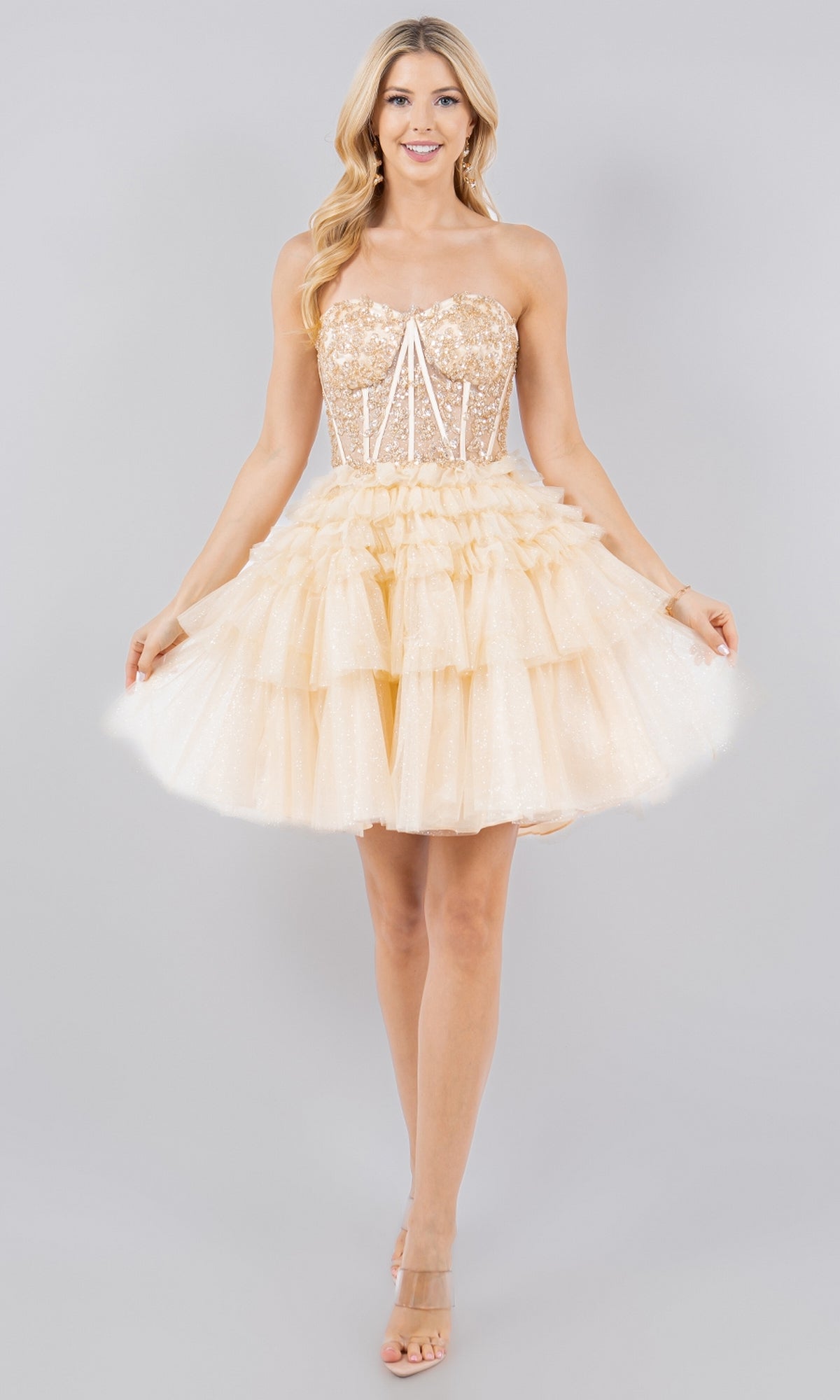 A woman models the Short Sweetheart Ruffled Homecoming Dress 5133J—a champagne sequin and tulle mini with a tiered ruffle skirt—channeling a perfect party look with nude heels against a plain backdrop.